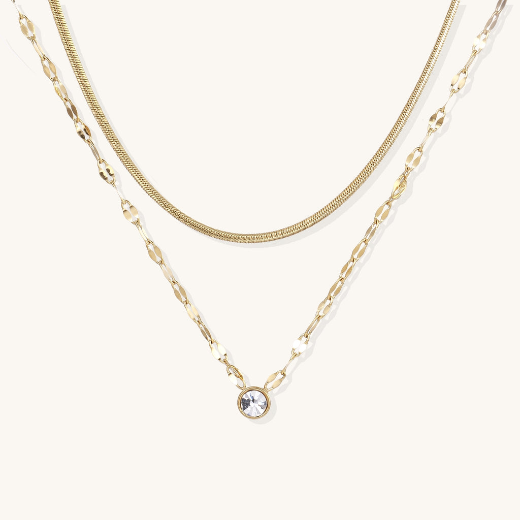 Two layered 14K gold plated necklaces - herringbone chain and delicate chain with circular crystal pendant on beige background.